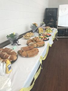 Long table, festively decorated, and filled with sandwiches, veggie trays with dip, chips, and fruit.